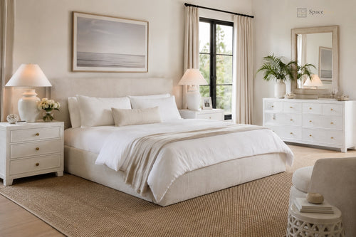 A beautifully styled bedroom featuring a low-profile platform bed dressed in white linens and a beige throw blanket, flanked by matching white nightstands with lamps. The room has a large woven jute rug, a white dresser with a mirror, and a window with sheer curtains.