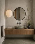 A modern floating bathroom vanity with a slatted wood cabinet, a stone vessel sink, a round black-framed mirror, and a hanging paper lantern pendant light in a minimalist bathroom setting.