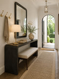 A styled entryway featuring a dark wood console table with a matching upholstered bench, a large black-framed mirror, a table lamp, wall hooks, and a long beige rug leading toward an arched doorway with outdoor greenery visible.