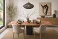 A styled dining room scene featuring a rectangular wooden dining table with a dark pedestal base, four upholstered dining chairs, a woven pendant light overhead, and a modern sideboard in the background with framed abstract wall art.