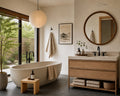 A modern bathroom interior with a freestanding oval bathtub, a light wood vanity with a stone countertop, a round wood-framed mirror, black faucets, folded towels, a small wooden side table, and a large window looking out onto greenery.
