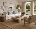 A bright, modern living room featuring a white sofa with light blue pillows, a light wood coffee table, a woven rattan armchair, and a large beige jute rug. Large windows with beige curtains look out to a green garden.