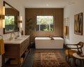A luxury modern bathroom interior featuring a double-sink vanity in dark wood, a white freestanding bathtub by a window, a patterned brown rug, and warm wood-paneled walls.