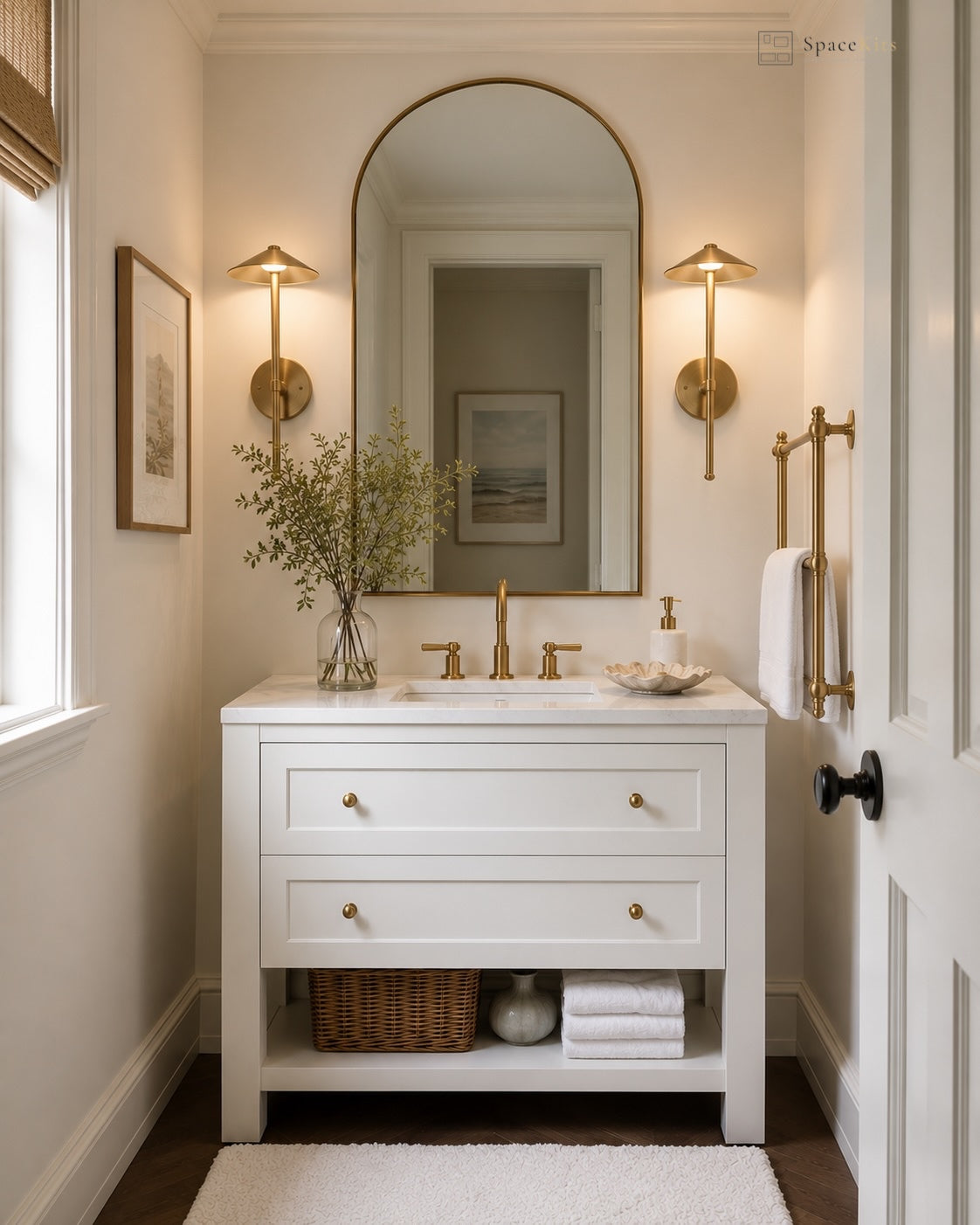 A white bathroom vanity cabinet with a white marble sink top, gold faucet and hardware, an arched gold-framed mirror, and matching gold sconces in a bright, neutral bathroom setting.