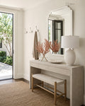 A styled entryway featuring a light wood console table with a matching bench, a large white wall mirror, and a white table lamp. A beige towel hangs from a wall hook, and a glass door on the left reveals greenery outside.