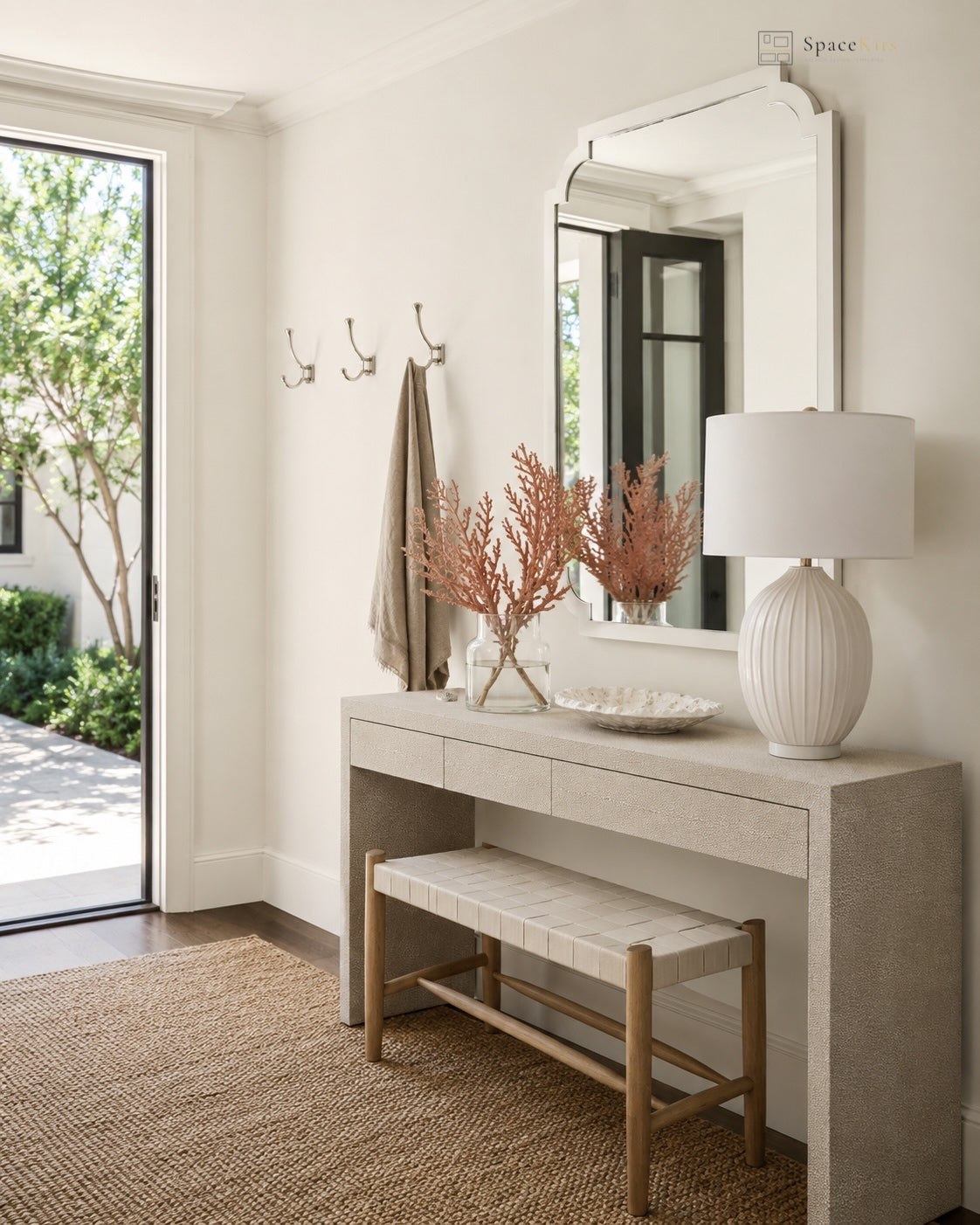 A styled entryway featuring a light wood console table with a matching bench, a large white wall mirror, and a white table lamp. A beige towel hangs from a wall hook, and a glass door on the left reveals greenery outside.