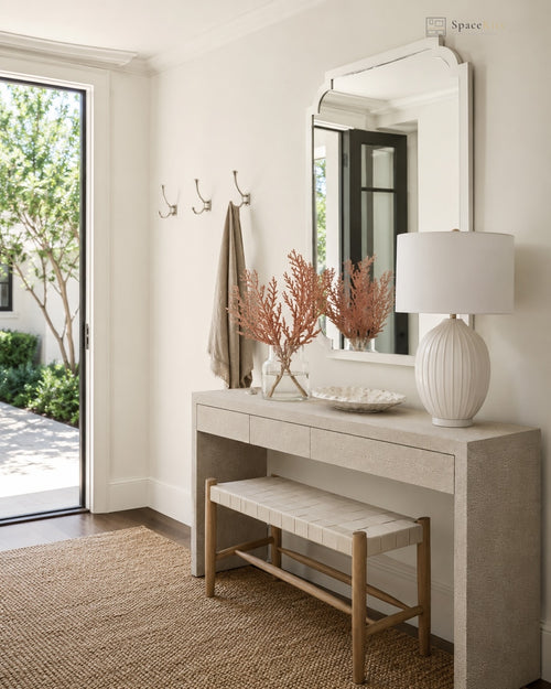A styled entryway featuring a light wood console table with a matching bench, a large white wall mirror, and a white table lamp. A beige towel hangs from a wall hook, and a glass door on the left reveals greenery outside.