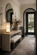 A modern entryway featuring a light wood console table with a matching upholstered bench, a large oval wall mirror, and a glowing cylindrical table lamp. A tall vase with dried branches sits on the table, and a textured area rug leads toward an arched black-framed glass door opening onto a garden.