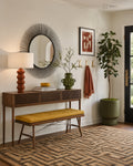 A styled entryway scene with a mid-century console table, yellow upholstered bench, round sunburst mirror, terracotta lamp, green ceramic vase, framed wall art, a rust-colored throw, and a tall potted plant.