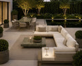Modern outdoor patio furniture set featuring a large U-shaped sectional sofa in off-white cushions with woven beige side panels, a matching rectangular coffee table with a green marble top, and a small side table with an illuminated lamp, arranged around a textured rug in front of a pool at dusk.