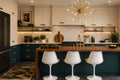 A modern kitchen interior with cream upper cabinets, dark teal lower cabinets, a wooden island, three white bar stools, gold hardware, and a gold starburst chandelier hanging above the island.