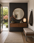 A modern entryway featuring a large round wood-framed mirror mounted on a dark charcoal wall above a floating wooden console. A woven rug, upholstered bench, and lit table lamp sit in the foreground, with an open door revealing a garden outside and a dark coat hanging on the wall.
