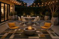 Outdoor patio furniture arrangement at dusk featuring two wire mesh armchairs with mustard yellow cushions facing a round concrete fire pit, with a white dining set and string lights in the background.