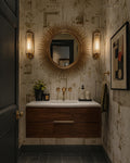 A styled bathroom interior featuring a dark wood floating vanity with a white sink, a round sunburst mirror, two gold wall sconces, patterned wallpaper, and framed wall art.