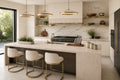 A modern kitchen island with a light beige stone countertop and base, three white bar stools with gold metal frames, and a marble backsplash. The kitchen features white cabinets, gold floating shelves, and three circular pendant lights.