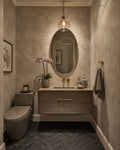 A modern bathroom vanity area with a wall-mounted wood sink cabinet, oval mirror, pendant light, dark herringbone tile floor, toilet, and a towel on the wall.