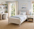 A light wood single bed dressed in white and blue bedding, styled in a bright, modern bedroom with a matching nightstand, desk, and a sailboat painting on the wall.