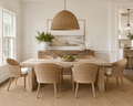 A dining room scene with a light wood rectangular table and six woven wicker chairs, under a large woven pendant light. The room has white walls, a wood sideboard with framed art and a plant, and a woven rug.