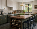 A modern kitchen interior with white upper cabinets, dark green lower cabinets and island, a wood-look countertop on the island, open shelving with bowls and cookware, bar stools, pendant lights, and a window looking out to greenery.