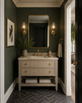 A styled bathroom interior featuring a cream ribbed vanity with a white marble countertop, gold faucets, a large gold-framed mirror, and dark green walls. A framed print, sconces, a plant, and folded towels are arranged around the vanity, with a doorway on the right showing a glimpse of another room.