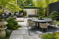 A beautifully styled modern Japanese-inspired outdoor patio featuring a round dark stone dining table with six woven wicker chairs, a dark gray sectional sofa, a stone lantern, a wooden raked path, and lush green bamboo and foliage.